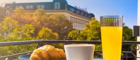 A table with a croissant, a cup, and a glass of orange juice, with a building and trees in the background.