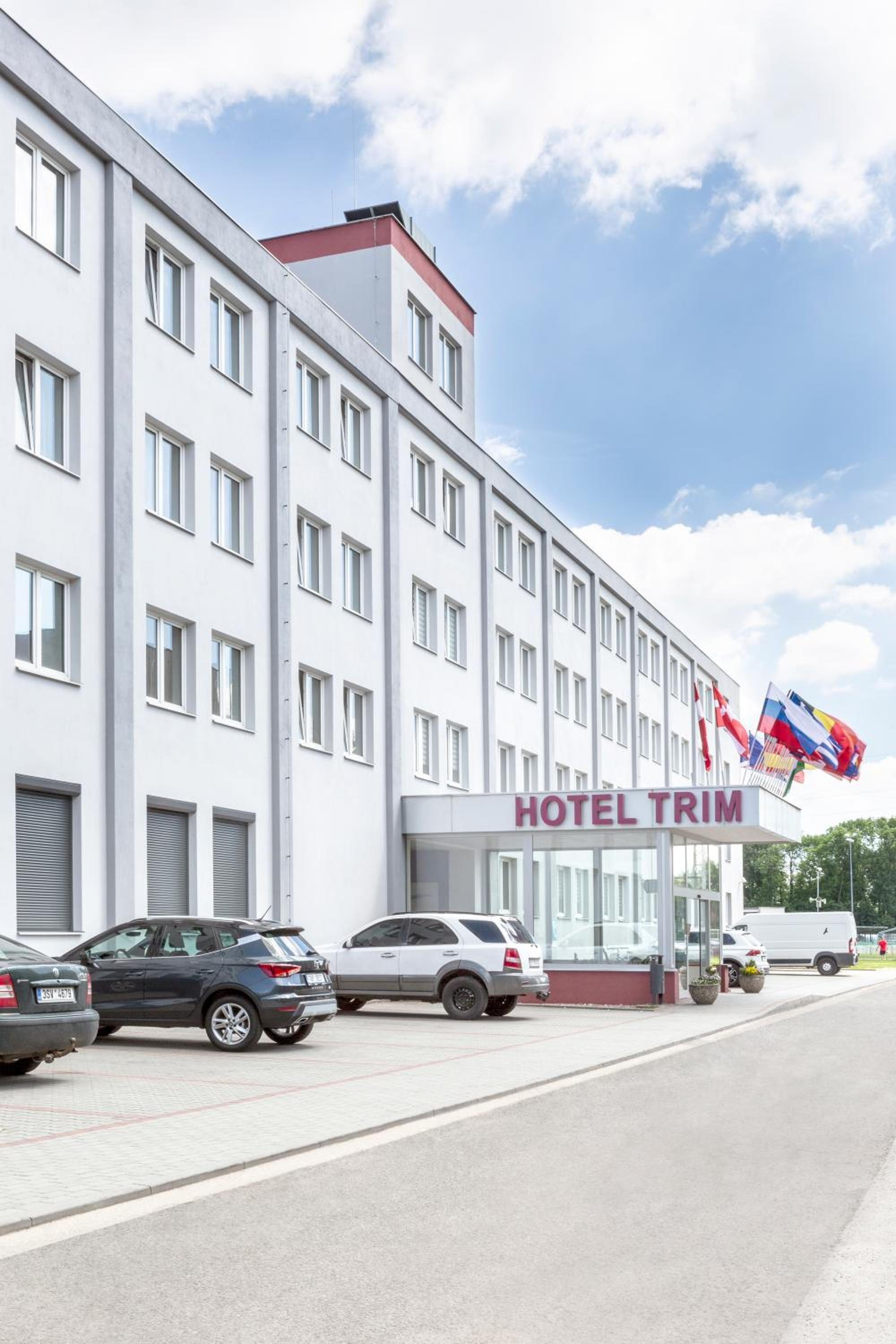 Un edificio de hotel de varios pisos con un letrero que dice 'HOTEL TRIM'. Hay coches estacionados al frente y el cielo está parcialmente nublado.