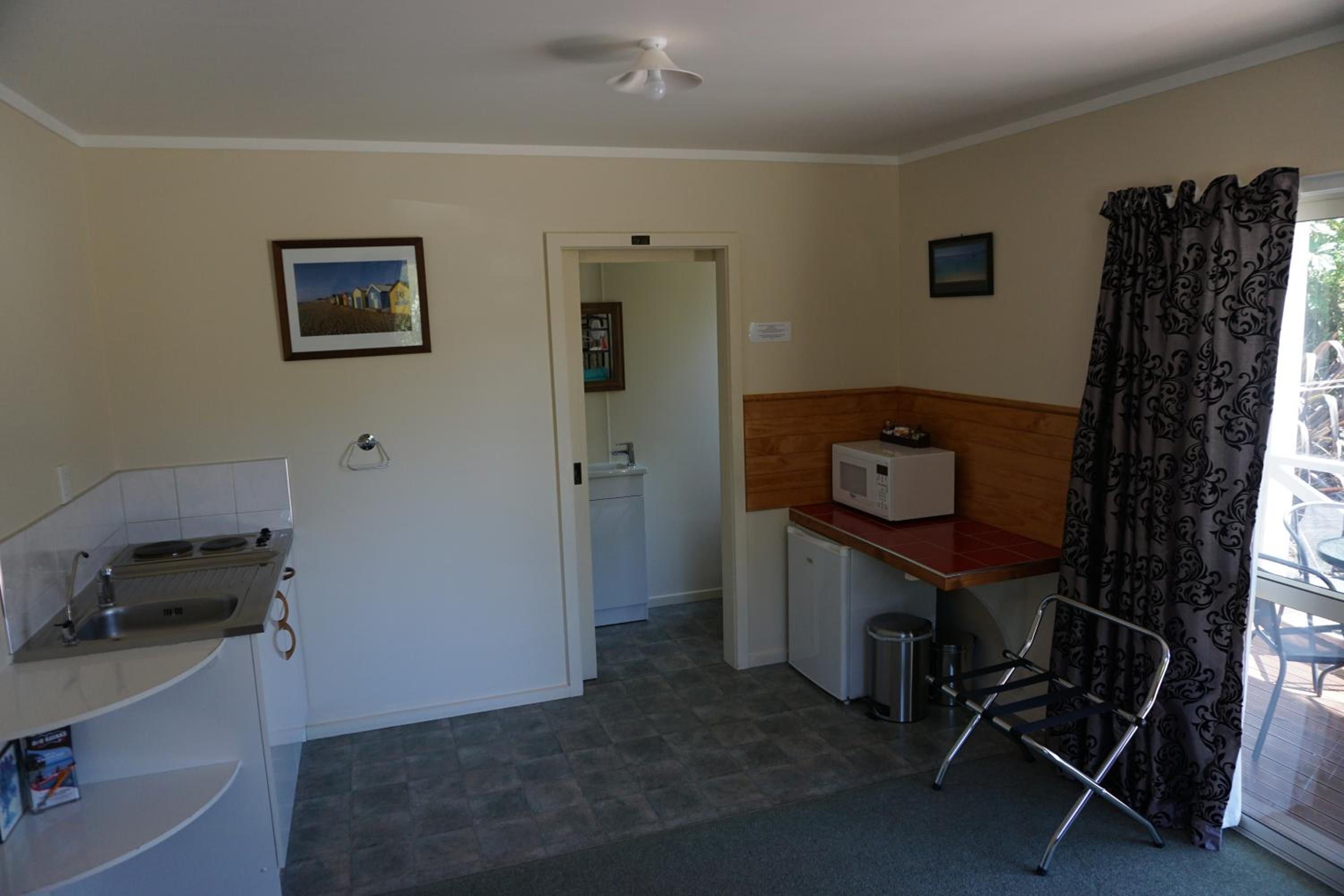 A compact kitchen with a microwave, sink, and a small refrigerator. There is a chair near the window and a door leading to another room.