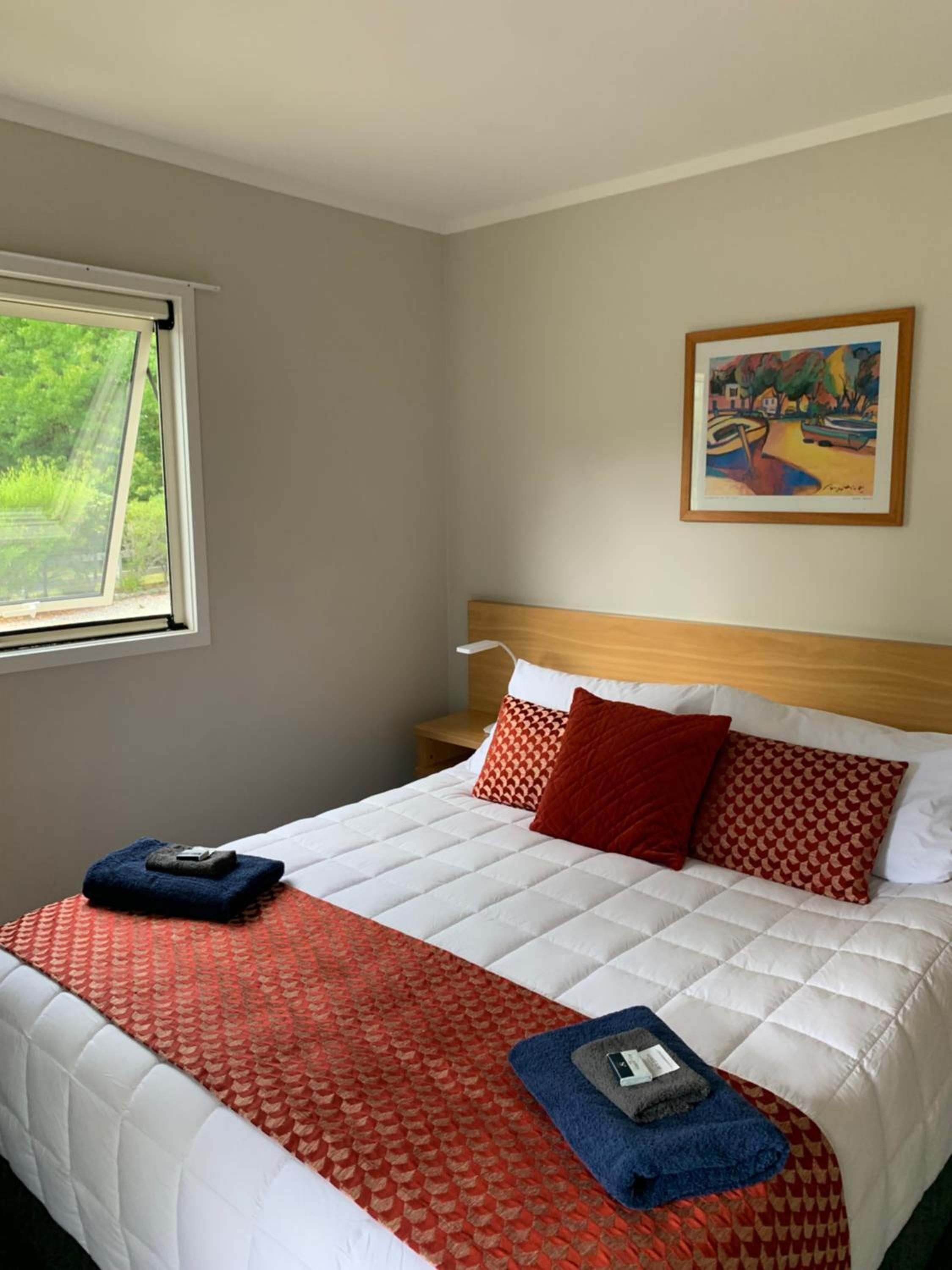 A hotel room with a bed, a painting on the wall, and a window with a view of greenery.