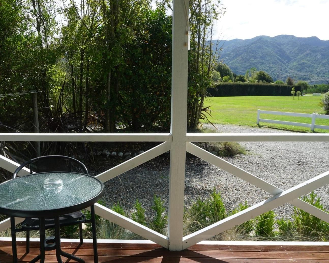 A glass table and chair on a wooden deck with a white railing, overlooking a grassy field and mountains.