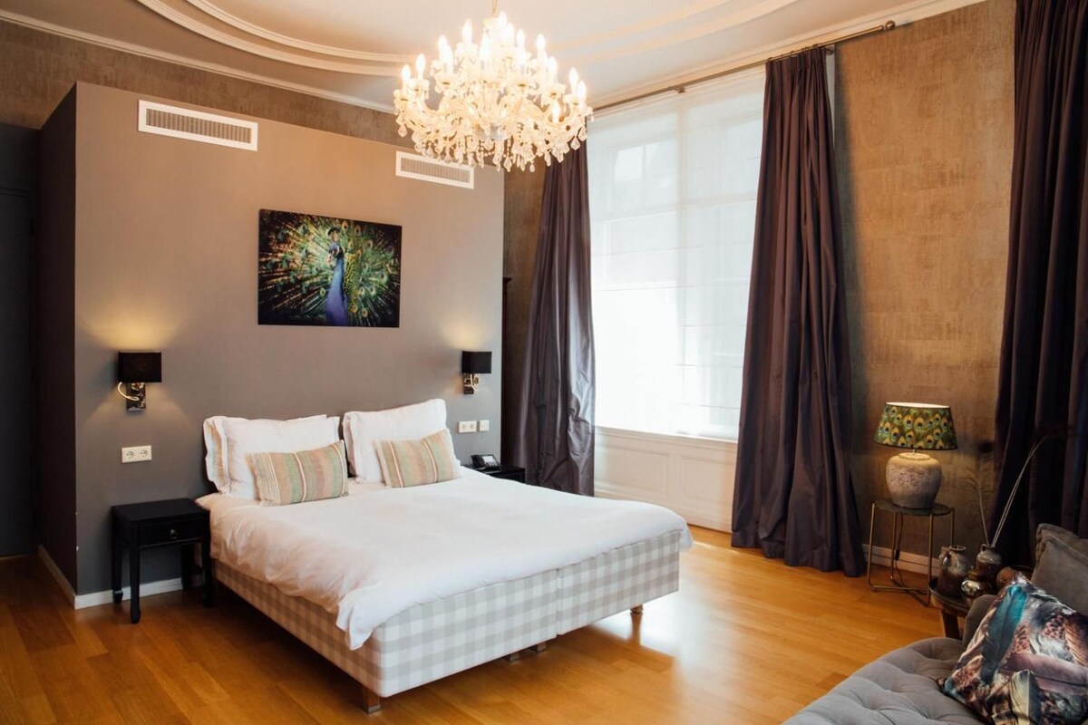 A bedroom with a large bed, a chandelier, a painting, and a window with curtains.
