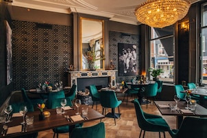 A restaurant interior with a chandelier, patterned wallpaper, and green upholstered chairs.