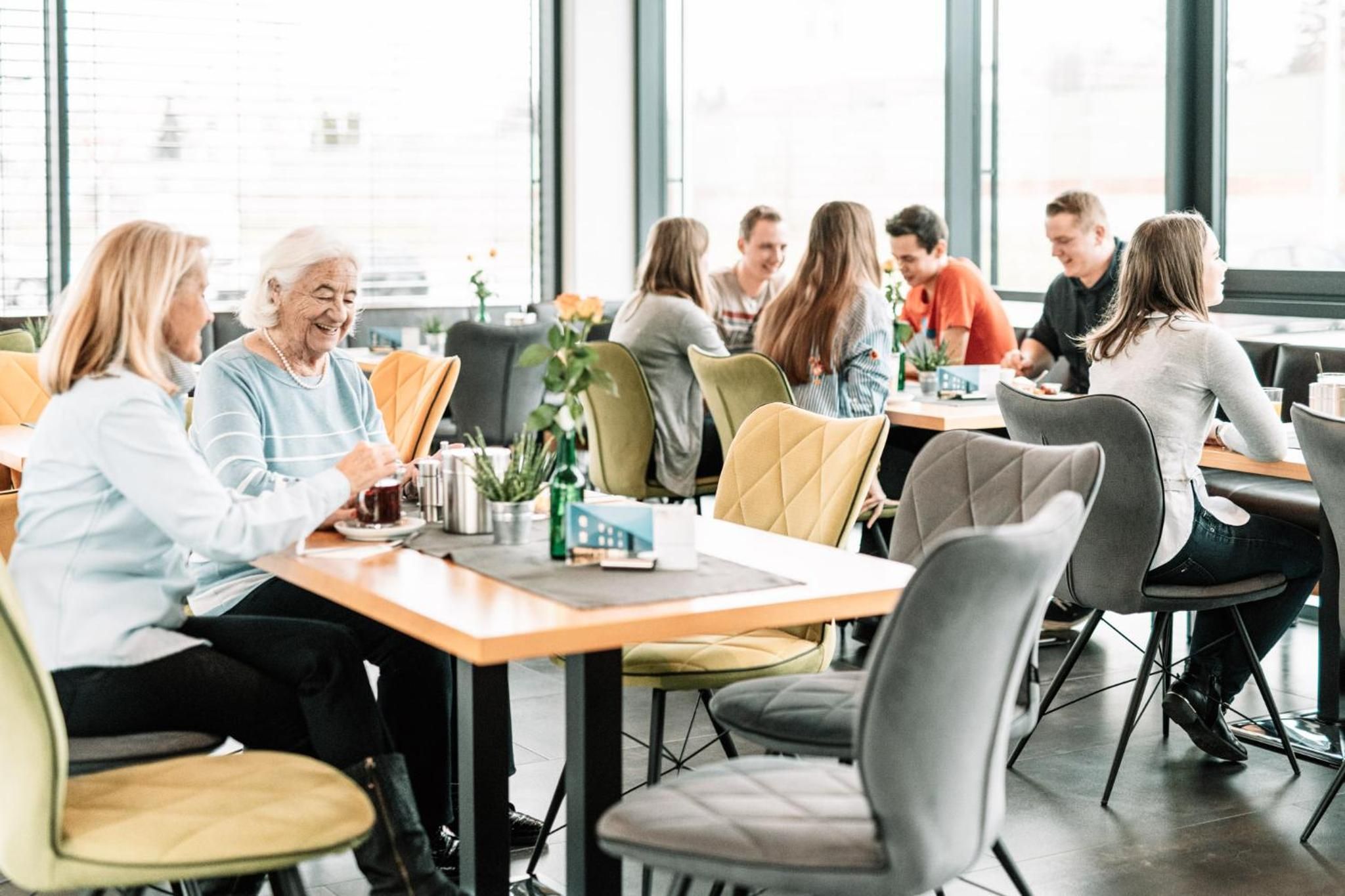 Eine Gruppe von Menschen sitzt an Tischen in einem hellen Innenraum mit großen Fenstern.