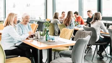 Eine Gruppe von Menschen sitzt an Tischen in einem hellen Innenraum mit großen Fenstern.