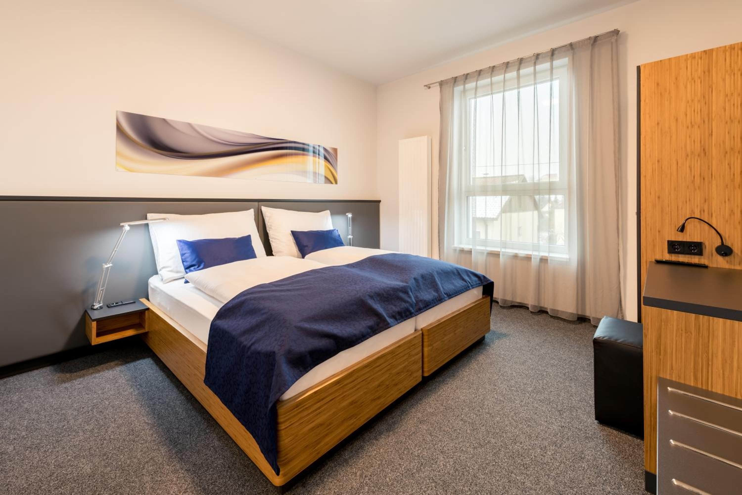 A hotel room with a bed, a bedside table, a lamp, a large window with curtains, and a wooden wardrobe.