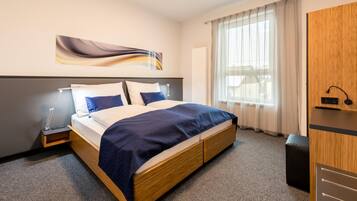 Ein Hotelzimmer mit Bett, Nachttisch, Lampe, großem Fenster mit Vorhängen und einem Holzschrank.