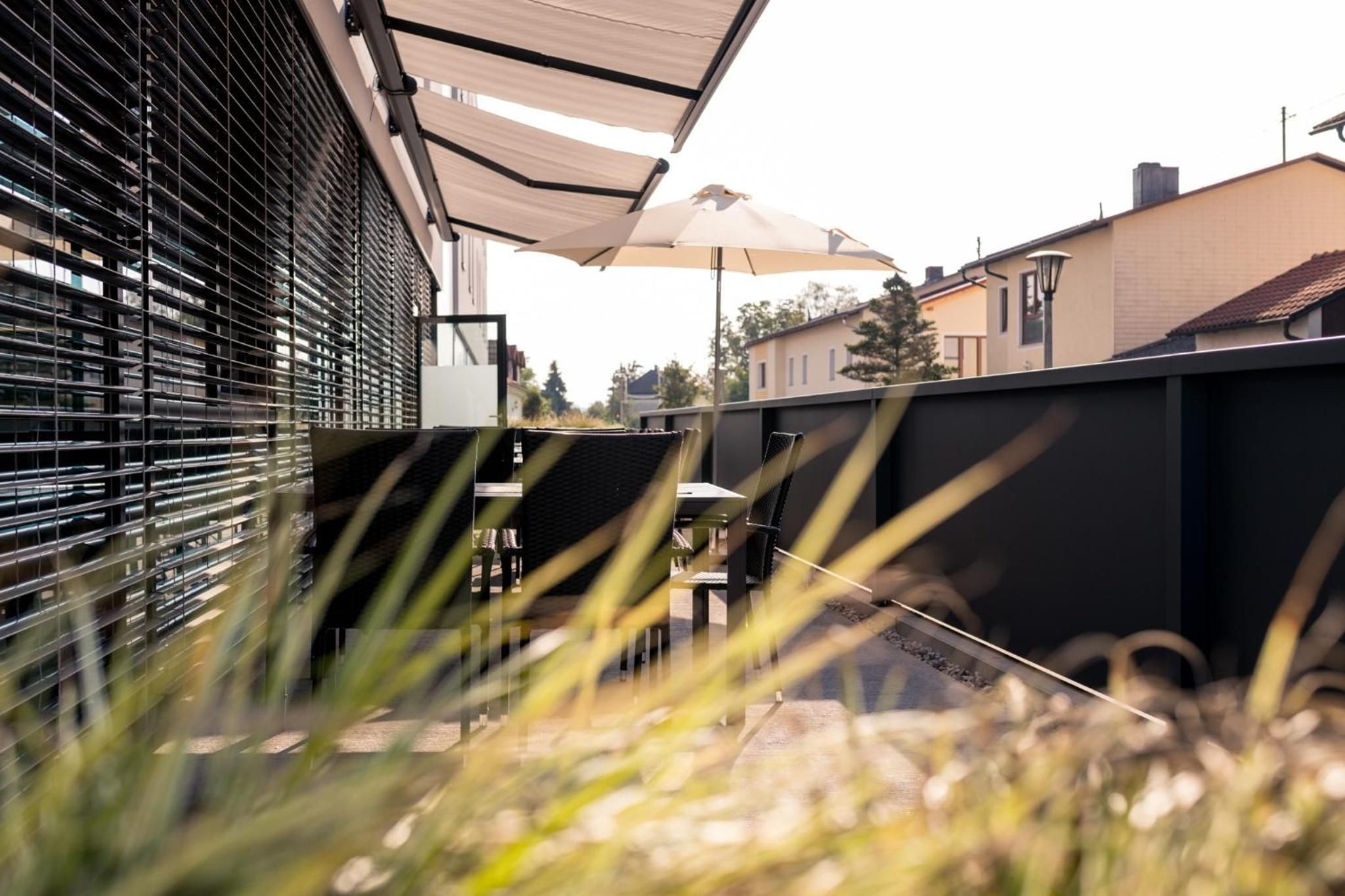 Ein Außenbereich mit Terrasse, Tisch und Stühlen unter einem Sonnensegel, umgeben von Zäunen und Bepflanzung.