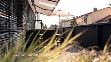 Ein Außenbereich mit Terrasse, Tisch und Stühlen unter einem Sonnensegel, umgeben von Zäunen und Bepflanzung.