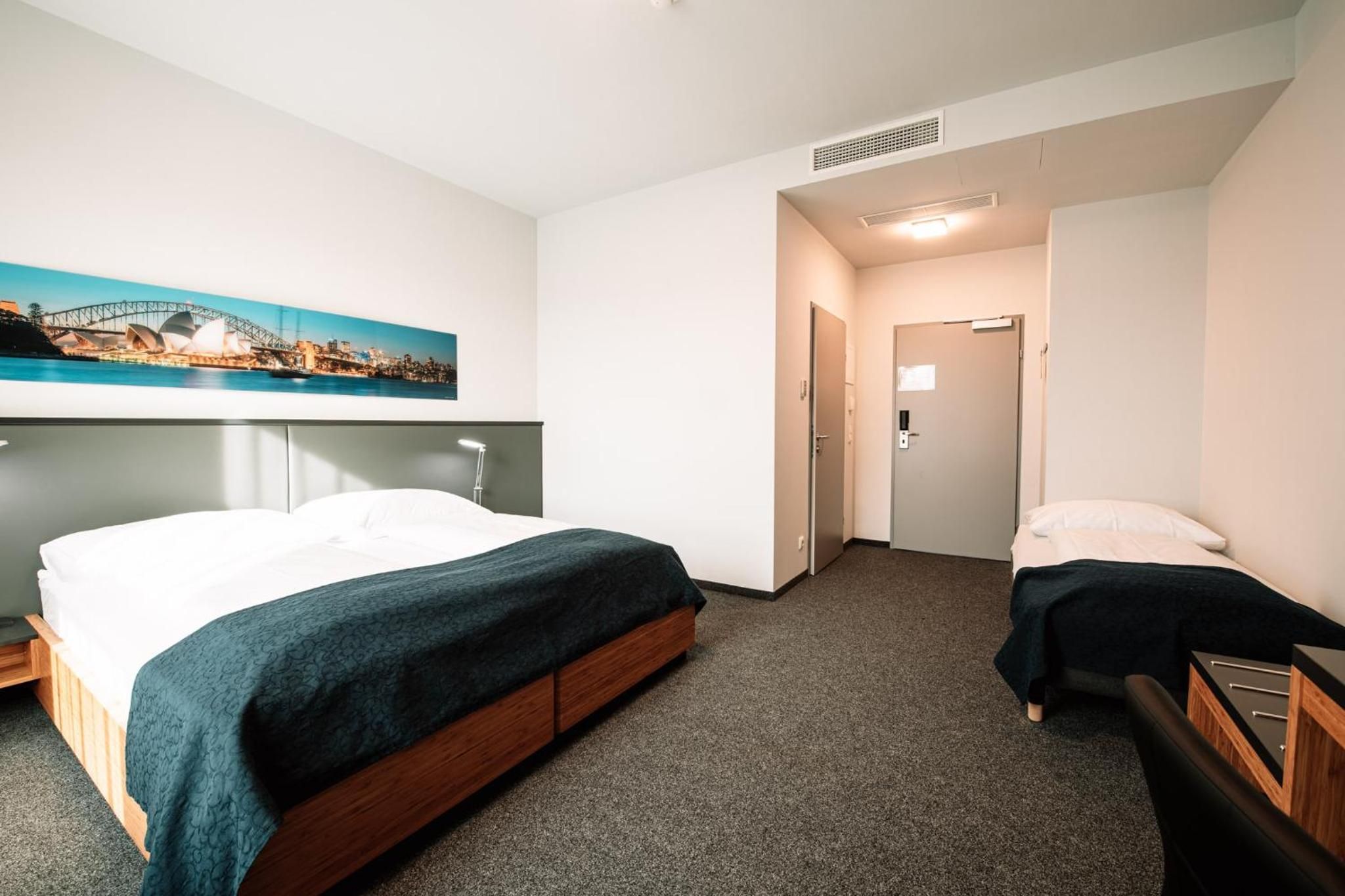 A hotel room with a bed, a bedside table, a chair, a desk, and a painting of a cityscape.