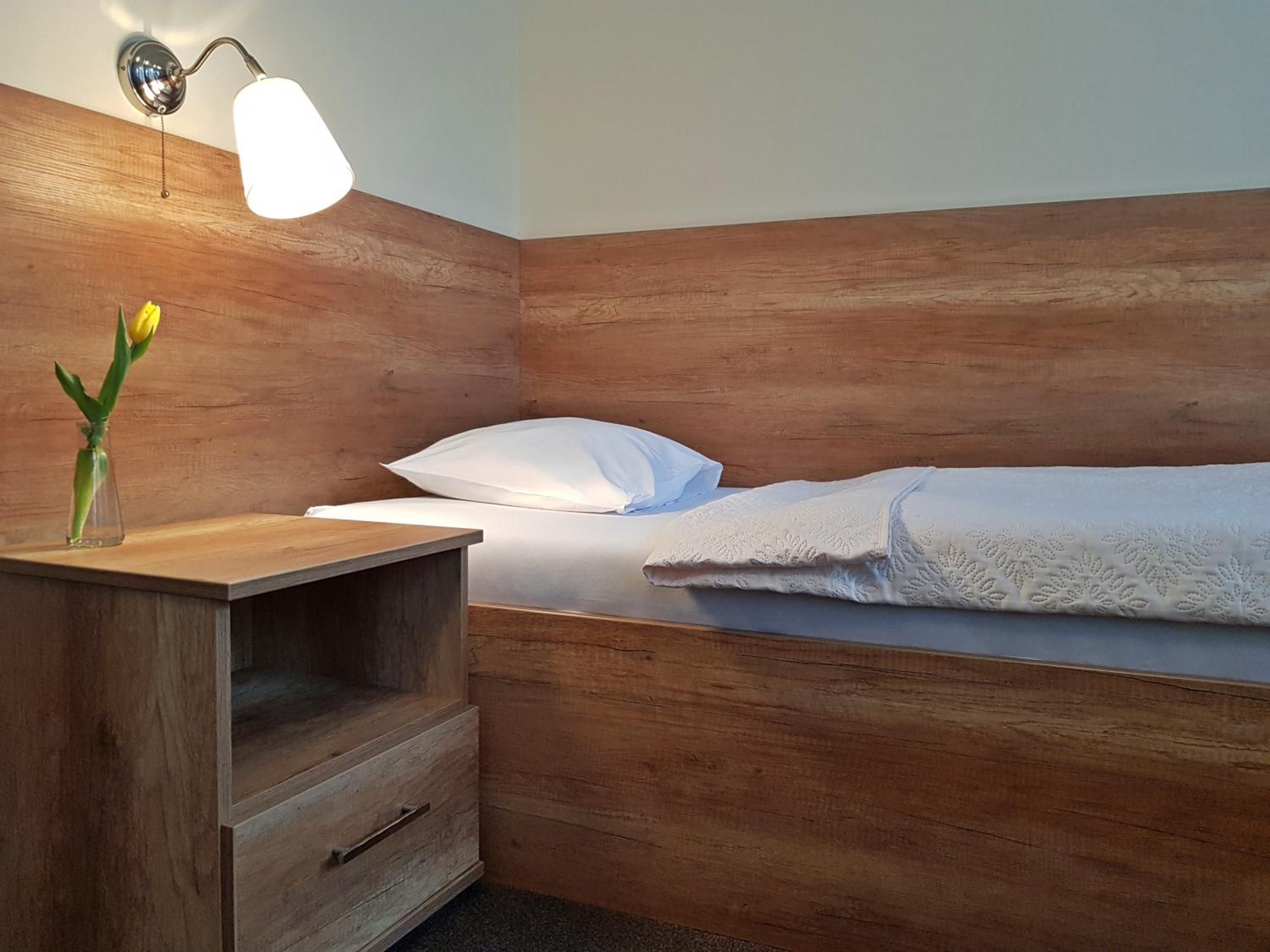 A single bed with a wooden headboard, a bedside table with a lamp, and a small vase with a flower.