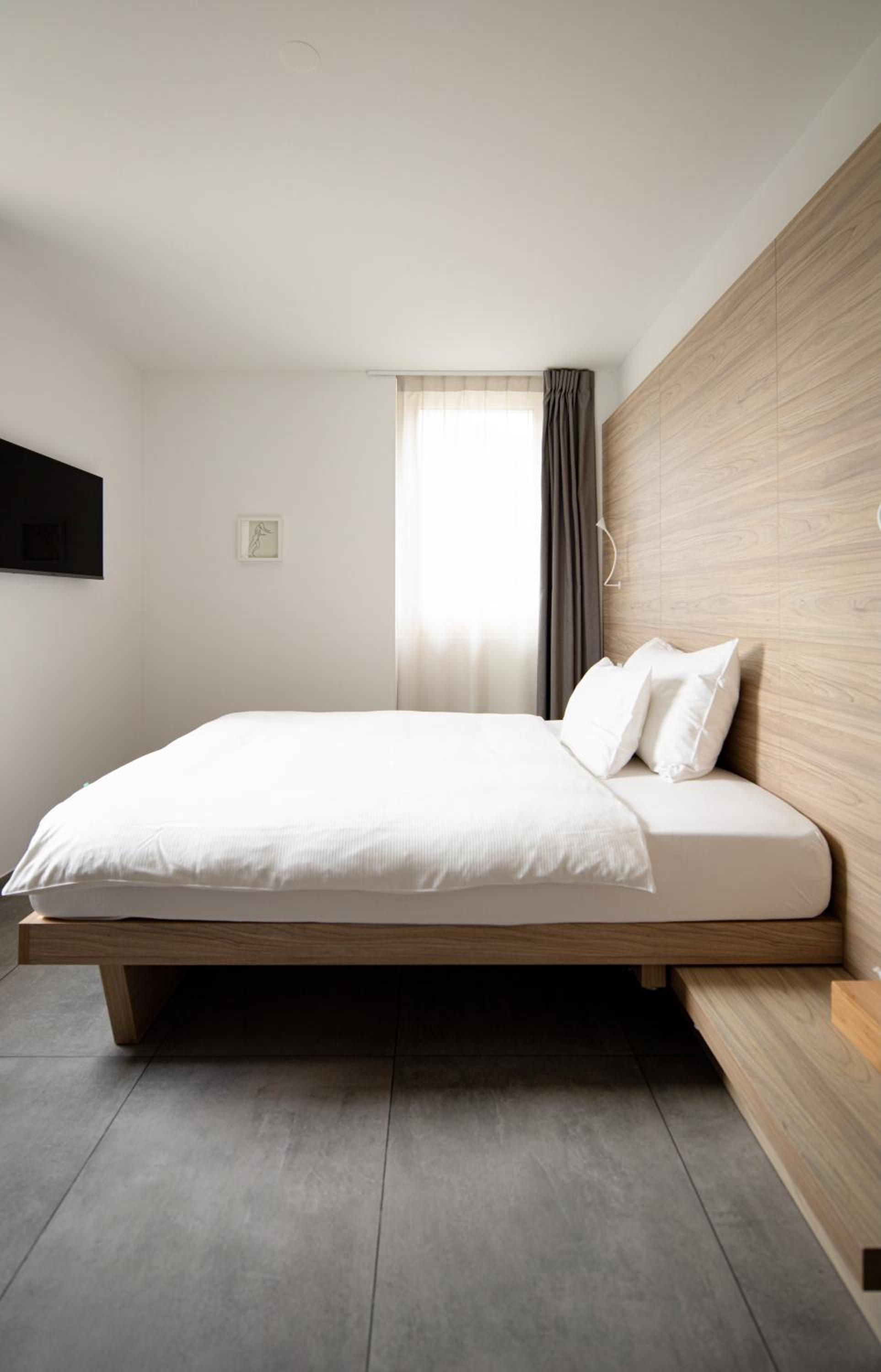 A modern bedroom with a minimalist design, featuring a bed with white bedding, a wooden headboard, and a large window with curtains.