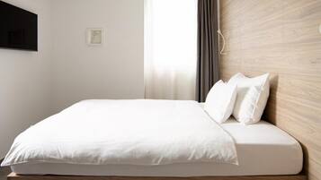 A modern bedroom with a minimalist design, featuring a bed with white bedding, a wooden headboard, and a large window with curtains.