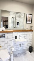Un baño con pared de azulejos blancos, encimera de madera, lavamanos, espejo, un cuadro enmarcado y un perchero para toallas.