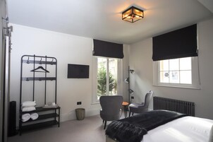 Habitación de hotel con cama, una silla, una mesita, radiador, ventana con vistas, televisor montado en la pared y un perchero con toallas.