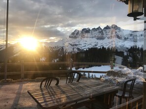 Una mesa y sillas de madera en un balcón con vista a montañas nevadas y un sol poniéndose al fondo.