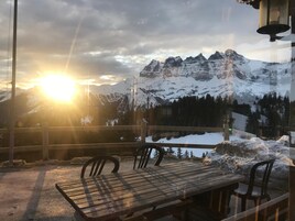 Una mesa y sillas de madera en un balcón con vista a montañas nevadas y un sol poniéndose al fondo.