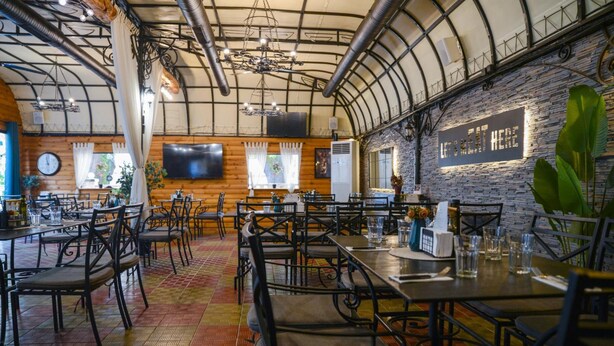 Ein Restaurant mit hoher Decke, freiliegenden Lüftungsrohren und einer Steinwand mit dem Schriftzug 'Let's Eat Here'. Tische mit Stühlen sind gedeckt, und ein Flachbildfernseher ist an der Wand befestigt.