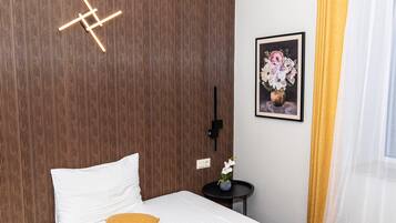 A hotel room with a bed, a wall-mounted light fixture, a framed floral artwork, and a small table with a vase.