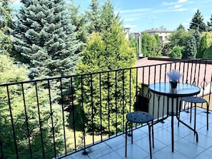 A balcony with a round table and two chairs, surrounded by a metal railing, overlooking a garden with various trees and a building in the background. - Pokoje Rezydencja Parysa (Lublin)