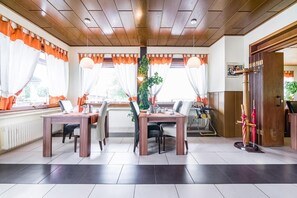 Een eetgedeelte met houten tafels en stoelen, grote ramen met oranje gordijnen, en een plant als decoratie.