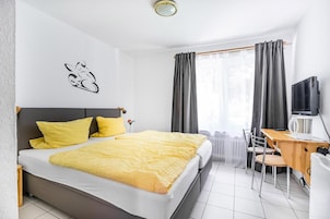 Habitación de hotel con cama, escritorio, silla, televisor y ventana con cortinas.