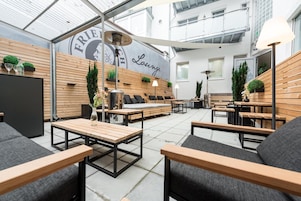 Un área de estar al aire libre con mobiliario de madera, una pared con la palabra 'Lounge' y un logotipo, y un techo con tragaluces.