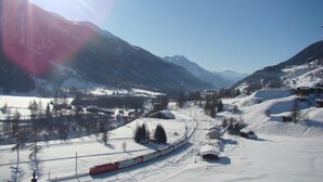 A snow-covered landscape with a train, mountains, and sparse buildings. - Hotel Weisshorn (Goms)