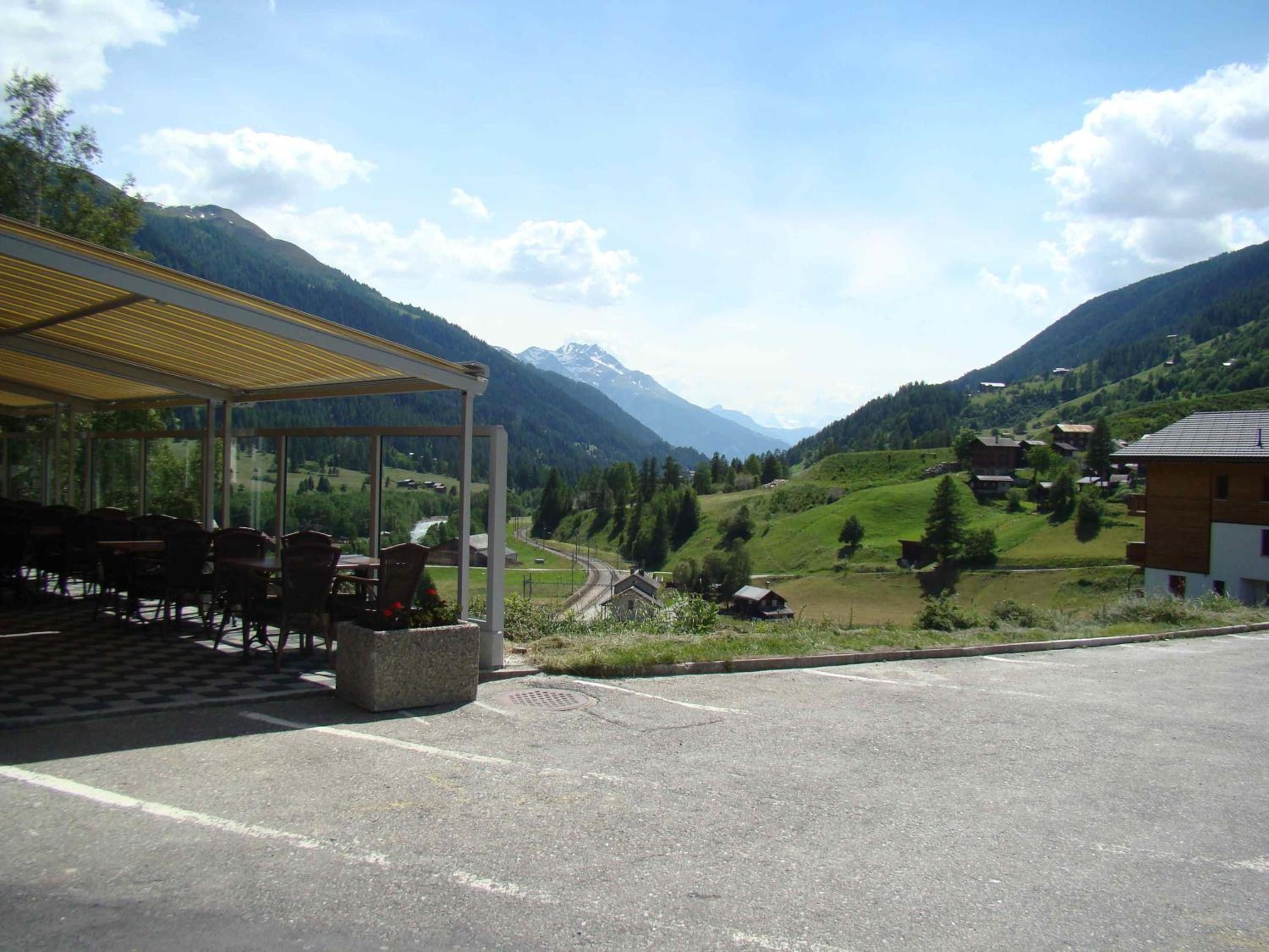 A covered outdoor seating area with tables and chairs, a paved road in the foreground, and a mountainous landscape with houses in the background.