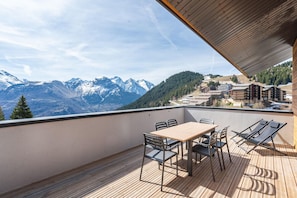 Uma varanda com mesa e cadeiras de madeira, com vista para uma paisagem montanhosa e edifícios ao longe.