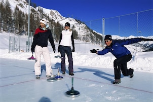 A snowy outdoor area with a fence, a person in a blue jacket throwing a curling stone, and another person in a white jacket observing.