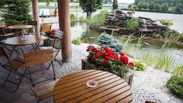 An outdoor seating area with wooden tables and chairs, a pond with a waterfall, and a view of a river.