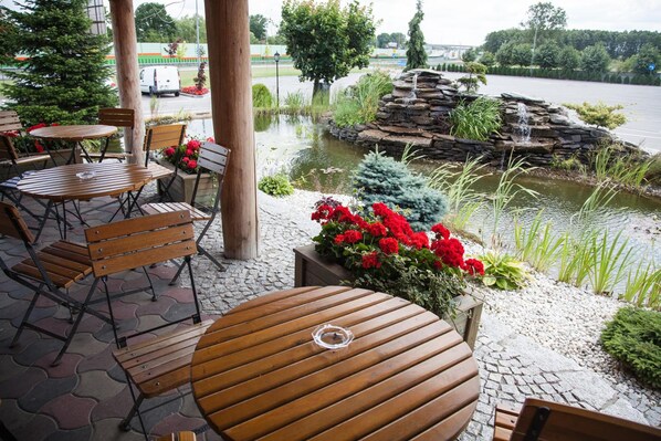 An outdoor seating area with wooden tables and chairs, a pond with a waterfall, and a view of a river.