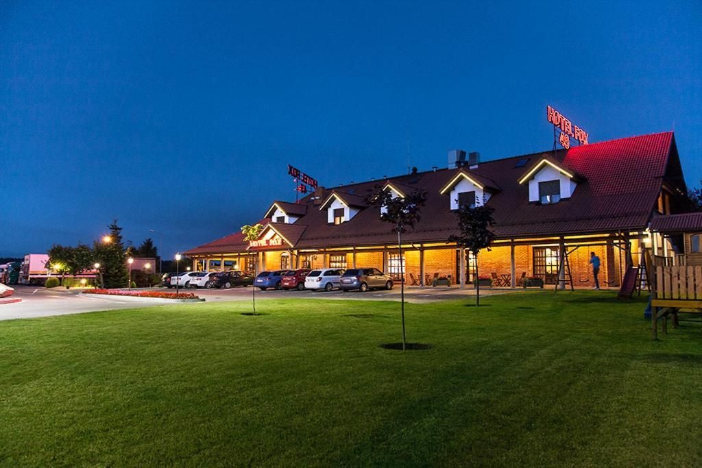 A hotel with a red roof and multiple windows, a parking lot with several cars, and a well-maintained lawn.