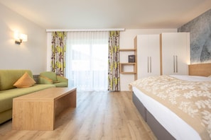 Habitación de hotel moderna con suelo de madera, un sofá verde, una mesa de centro de madera, una cama con colcha estampada, un armario y un gran ventanal con cortinas.