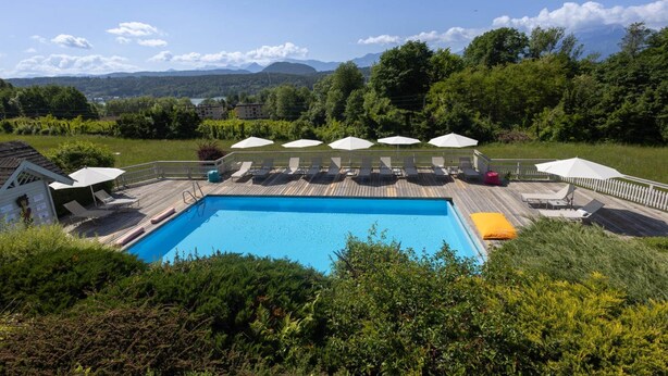 Una piscina rectangular con una terraza de madera, rodeada de sillas de descanso y sombrillas, con montañas y árboles al fondo.