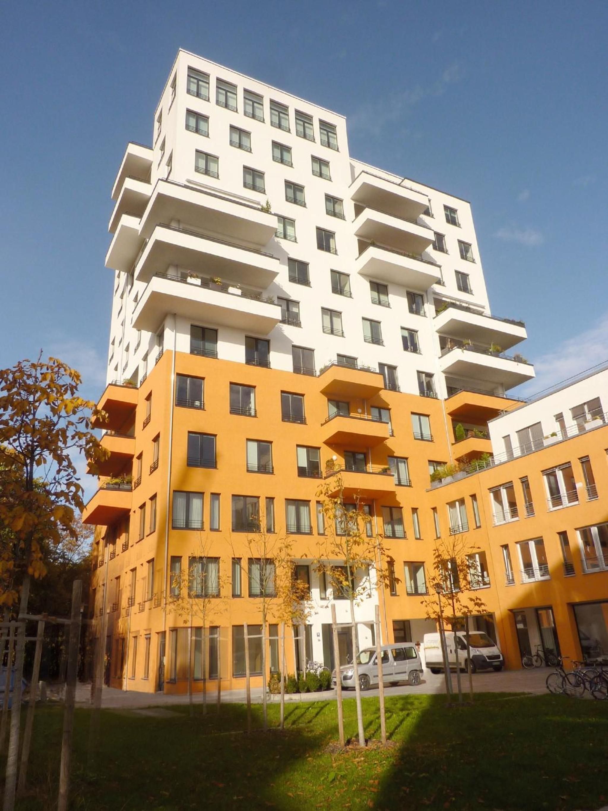 A multi-story building with balconies, a flat roof, and a parking area with cars and bicycles.