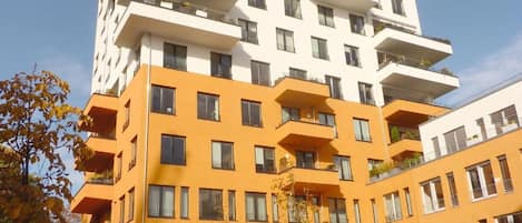 Un immeuble à plusieurs étages doté de balcons, d’un toit plat et d’un espace de stationnement où sont garées des voitures et des vélos.