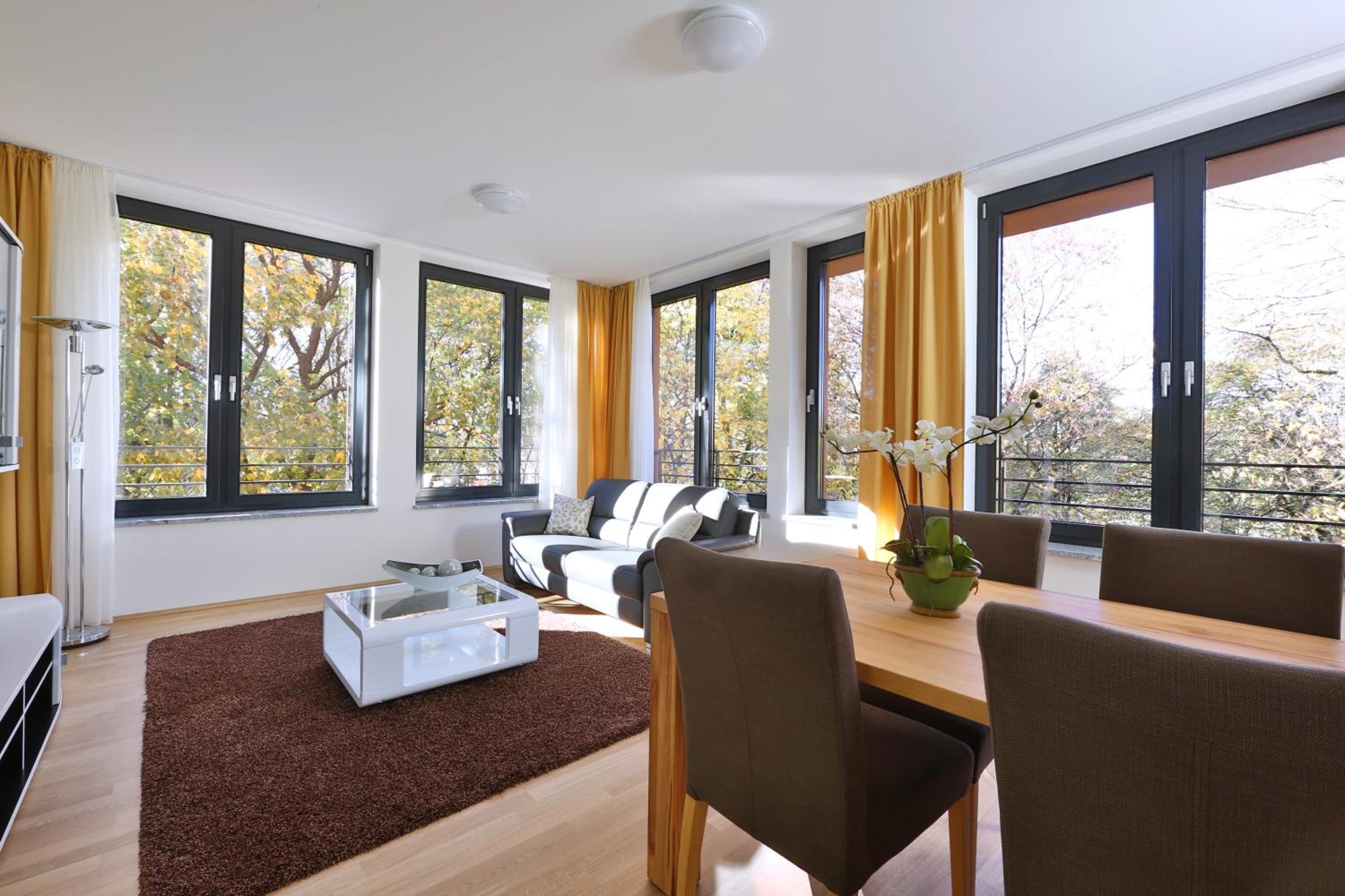 Un salon moderne avec un canapé, une table à manger et des chaises. De grandes fenêtres sont ornées de rideaux, et une plante est posée sur la table.