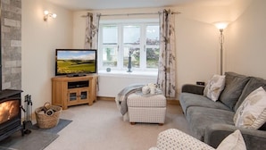 A cozy living room with a fireplace, a television, a sofa, and a window with curtains. - Chatton Cottage (Alnwick)