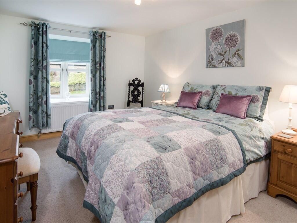 A bedroom with a bed, a nightstand, a chair, a window with curtains, and a wall-mounted lamp.