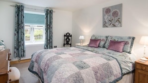A bedroom with a bed, a nightstand, a chair, a window with curtains, and a wall-mounted lamp. - Chatton Cottage (Alnwick)