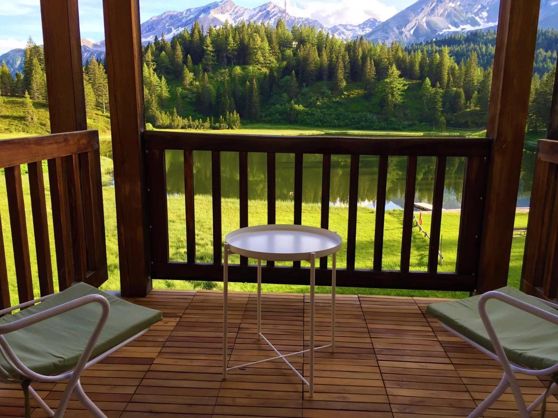 A wooden deck with a table and two chairs, overlooking a lake and a forested mountain range.