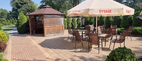An outdoor seating area with tables and chairs, a wooden structure, and a large umbrella with the brand 'TYSKIE' visible.