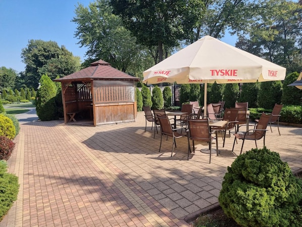 An outdoor seating area with tables and chairs, a wooden structure, and a large umbrella with the brand 'TYSKIE' visible.