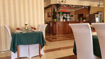 A hotel dining area with a green table set for four, white chairs, a bar area with a Coca-Cola refrigerator, and a patterned wallpaper background.