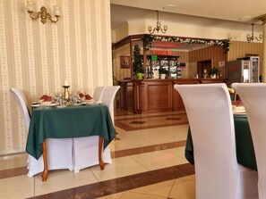 A hotel dining area with a green table set for four, white chairs, a bar area with a Coca-Cola refrigerator, and a patterned wallpaper background.