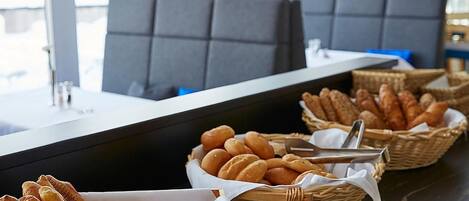 Zona de buffet con pan y pasteles en cestas, una barra y asientos.