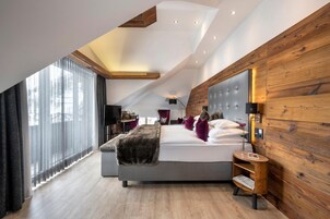 A modern bedroom with a sloped ceiling, a large bed, a wooden accent wall, and a view through a large window.