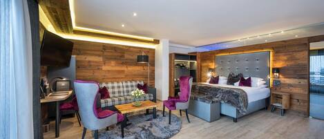 A modern hotel room with a wooden accent wall, a sofa, a chair, a bed, and a desk with a lamp.